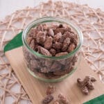 Ein Glasgefäß mit gebrannten Mandeln aus der Heißluftfritteuse steht auf einem Holzbrett auf einem geflochtenen Tischset, daneben liegen ein paar gezuckerte Mandeln.