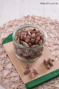 Ein Glas gebrannte Mandeln aus der Heißluftfritteuse mit ein paar losen Mandeln auf einem Holzbrett.