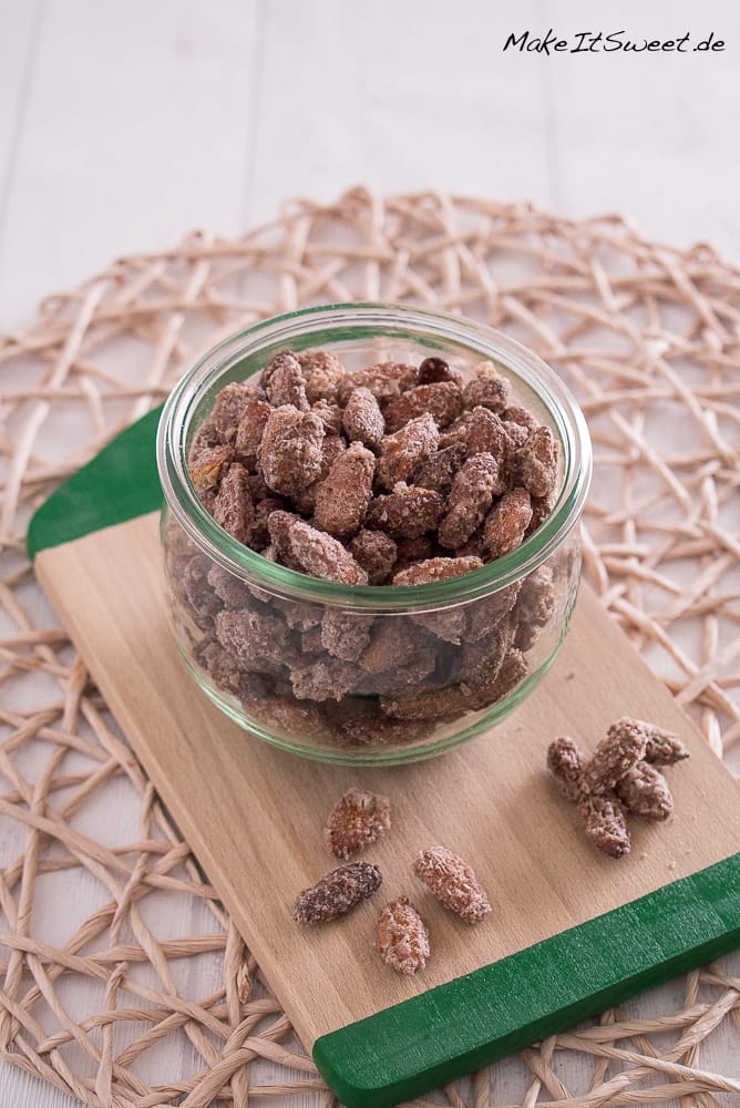 Gebrannte Mandeln aus der Heißluftfritteuse 1 Ein Glasgefäß mit gebrannten Mandeln aus der Heißluftfritteuse steht auf einem Holzbrett auf einem geflochtenen Tischset, daneben liegen ein paar gezuckerte Mandeln.