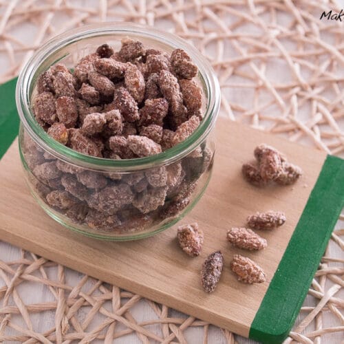 Gebrannte Mandeln in einem Glasgefäß auf einem Holzbrett, mit zusätzlichen Gebrannten Mandeln aus der Heißluftfritteuse in der Nähe.