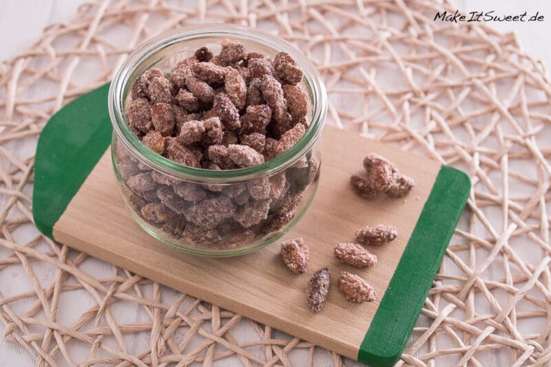 Gebrannte Mandeln in einem Glasgefäß auf einem Holzbrett, mit zusätzlichen Gebrannten Mandeln aus der Heißluftfritteuse in der Nähe.