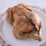 Ganzes gefülltes Hähnchen in der Heißluftfritteuse mit Maronen, Apfel und Zwiebeln Ein ganzes gefülltes Hähnchen mit gebräunter Haut liegt auf einem weißen Teller, der auf einem geflochtenen Tischset auf einer weißen Holzfläche platziert ist.