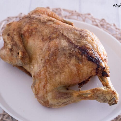 Ganzes gefülltes Hähnchen in der Heißluftfritteuse mit Maronen, Apfel und Zwiebeln 3 Ein ganzes gefülltes, goldbraun gebratenes Hähnchen liegt auf einem weißen Teller, der auf einem geflochtenen Tischset auf einer hellen Holzfläche steht.