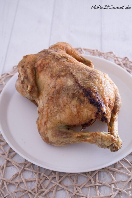 Ganzes gefülltes Hähnchen in der Heißluftfritteuse mit Maronen, Apfel und Zwiebeln 2 Ein ganzes gefülltes Hähnchen mit gebräunter Haut liegt auf einem weißen Teller, der auf einem geflochtenen Tischset auf einer weißen Holzfläche platziert ist.