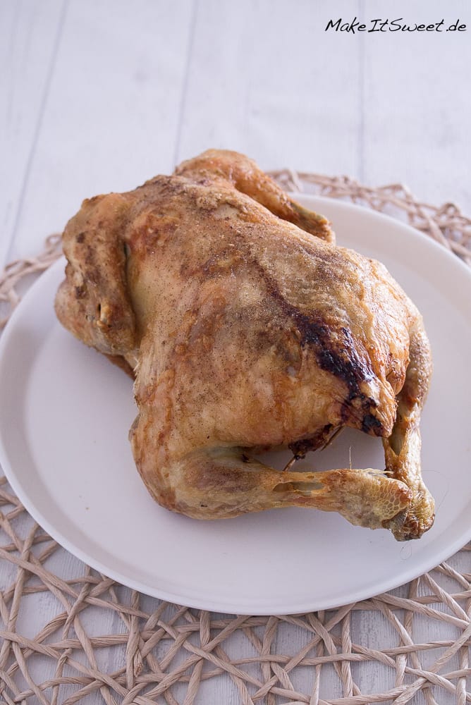 Ganzes gefülltes Hähnchen in der Heißluftfritteuse mit Maronen, Apfel und Zwiebeln 1 Ein ganzes gefülltes Hähnchen mit gebräunter Haut liegt auf einem weißen Teller, der auf einem geflochtenen Tischset auf einer weißen Holzfläche platziert ist.