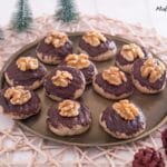 Ein rundes Tablett enthält Plätzchen, die mit Schokolade und Walnusshälften belegt sind. Das Tablett steht auf einer gewebten Matte, daneben stehen kleine künstliche Tannenbäume und ein Tannenzapfen. Der Hintergrund ist hell und neutral.