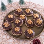 Walnuss-Zimt-Plätzchen Ein rundes Tablett mit elf Plätzchen, belegt mit Schokolade und Walnusshälften, steht auf einer gewebten Matte. Kleine künstliche Tannenbäume und zwei Tannenzapfen schmücken den weißen Holztisch rund um das Tablett.