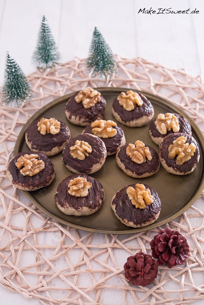 Walnuss-Zimt-Plätzchen 2 Ein rundes Tablett mit elf Plätzchen, belegt mit Schokolade und Walnusshälften, steht auf einer gewebten Matte. Kleine künstliche Tannenbäume und zwei Tannenzapfen schmücken den weißen Holztisch rund um das Tablett.