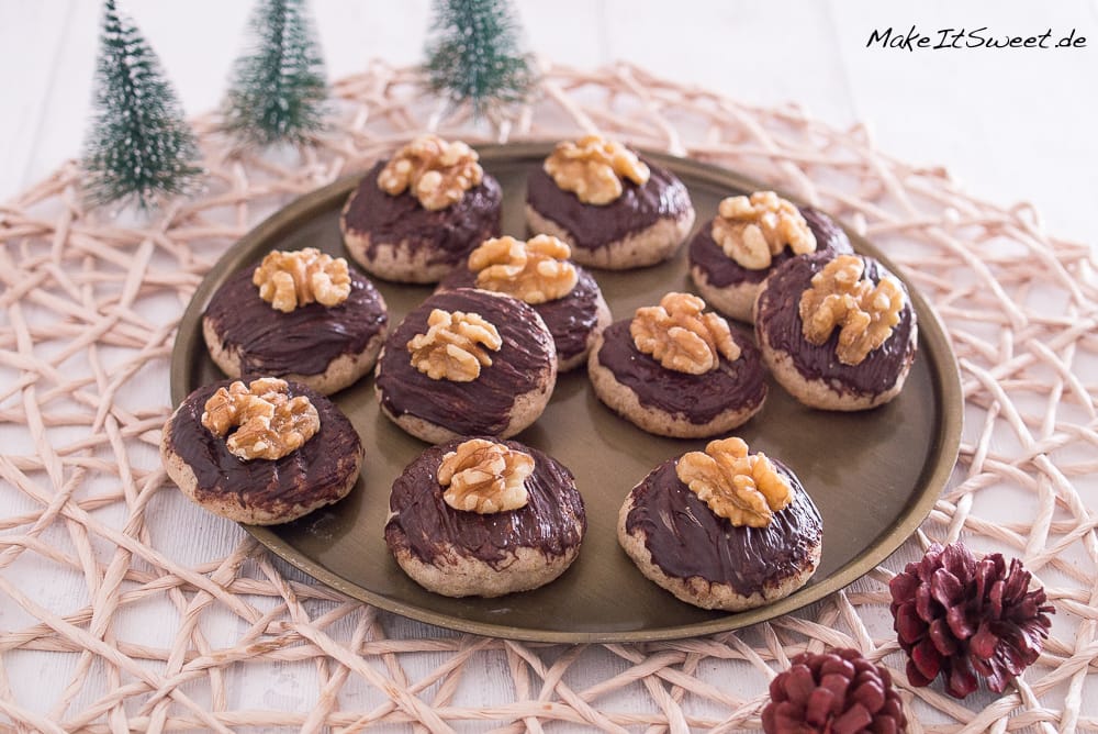 Runde Walnuss-Zimt-Plätzchen mit Schokolade und Walnusshälften auf einem Tablett angerichtet.