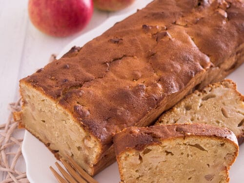 Apfel-Quark-Kuchen aus der Kastenform mit zwei geschnittenen Scheiben, einer Holzgabel und zwei Äpfeln auf einem weißen Teller.