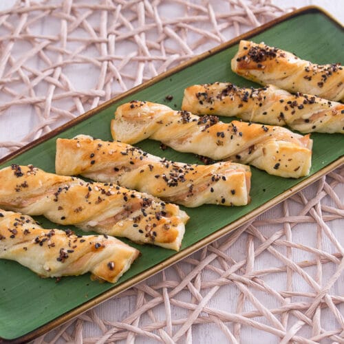 Sechs gedrehte Blätterteigstangen mit Sesam, Mohn und Räucherlachs darauf, nebeneinander auf einer grünen, rechteckigen Platte angeordnet.