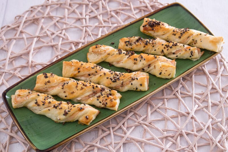 Sechs gedrehte Blätterteigstangen mit Sesam, Mohn und Räucherlachs darauf, nebeneinander auf einer grünen, rechteckigen Platte angeordnet.