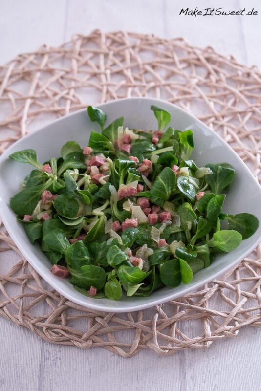 Feldsalat mit Speck und Brühe 2 Feldsalat mit Speck in einer weißen Schale, mit frischen grünen Salatblättern und Schinkenwürfeln.