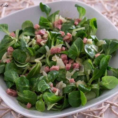 Feldsalat mit Speck und Brühe 3 Quadratische weiße Schale mit Feldsalat mit Speck, mit frischem Grün, Schinkenwürfeln und Zwiebeln.