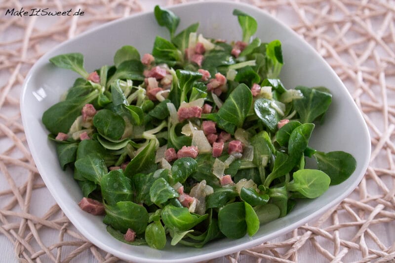 Feldsalat mit Speck und Brühe 1 Quadratische weiße Schale mit Feldsalat mit Speck, mit frischem Grün, Schinkenwürfeln und Zwiebeln.