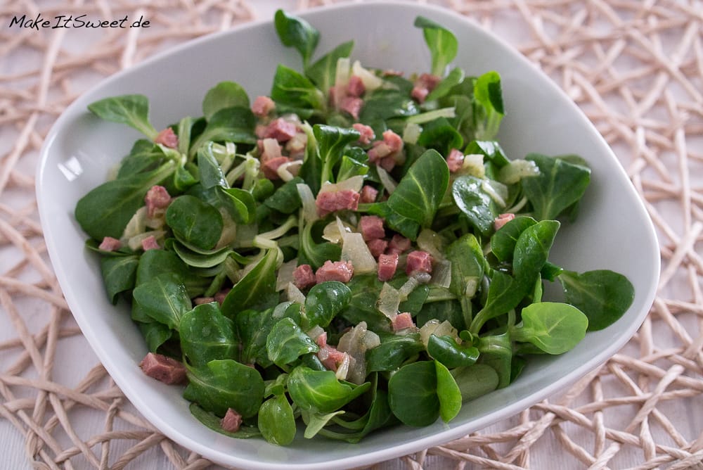 Quadratische weiße Schale mit Feldsalat mit Speck, mit frischem Grün, Schinkenwürfeln und Zwiebeln.