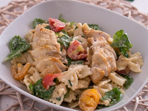 Nudelsalat mit gegrilltem Hähnchen, Tomaten, Rucola und Soße, serviert in einer weißen Schale.