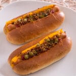 Zwei Hot Dogs mit Cheddar-Käse, Relish, Ketchup und knusprigen Röstzwiebeln auf einem weißen Teller.