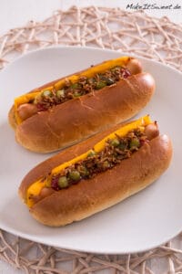 Zwei Hot Dogs mit Cheddar-Käse, Relish, Ketchup und knusprigen Röstzwiebeln auf einem weißen Teller.