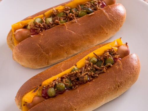 Zwei Hot Dogs mit Cheddar-Käse, Relish, Ketchup und knusprigen Röstzwiebeln auf einem weißen Teller.