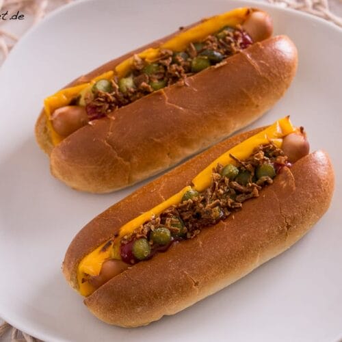 Zwei Hot Dogs auf einem weißen Teller, belegt mit Käse, Relish, Zwiebeln und Ketchup.