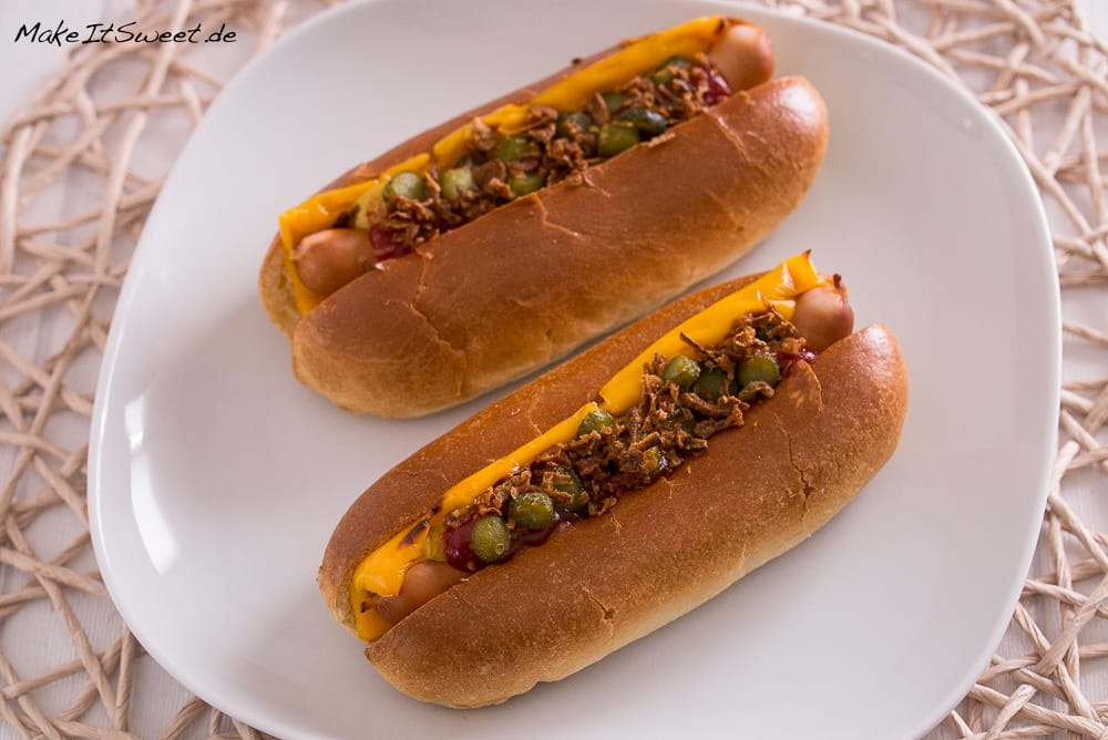 Zwei Hot Dogs auf einem weißen Teller, belegt mit Käse, Relish, Zwiebeln und Ketchup.
