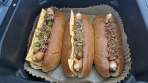 Drei Hot Dogs in Brötchen, belegt mit Käse, Ketchup, Essiggurken und knusprigen Zwiebeln auf Pergamentpapier in einem Tablett.