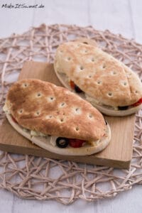 Zwei Panini-Fladenbrot-Sandwiches mit Oliven, Feta und Gemüse auf einem Holzbrett, knusprig und frisch aus einer Heißluftfritteuse.