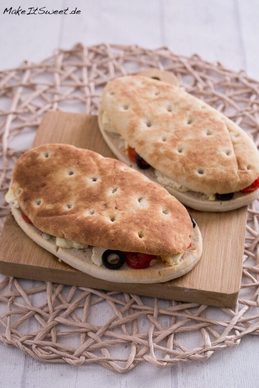 Zwei Panini-Fladenbrot-Sandwiches mit Oliven, Feta und Gemüse auf einem Holzbrett, knusprig und frisch aus einer Heißluftfritteuse.
