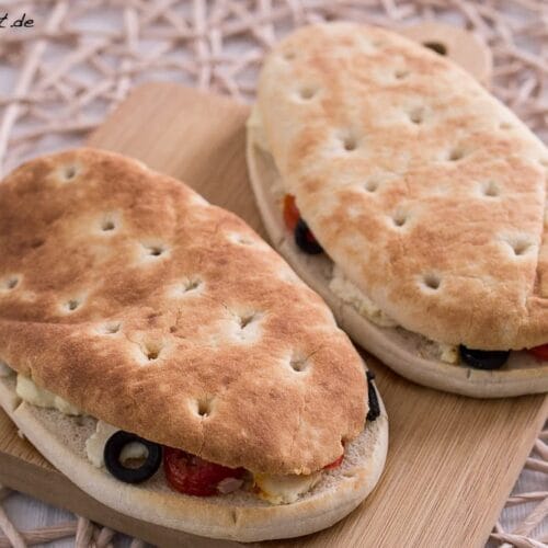 Zwei ovale Fladenbrote, belegt mit Feta, Oliven und Tomaten auf einem Holzbrett.