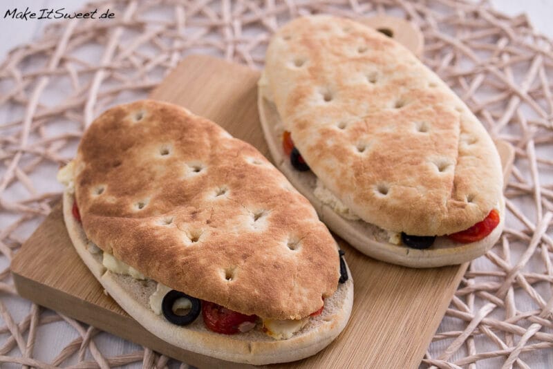 Zwei ovale Fladenbrote, belegt mit Feta, Oliven und Tomaten auf einem Holzbrett.