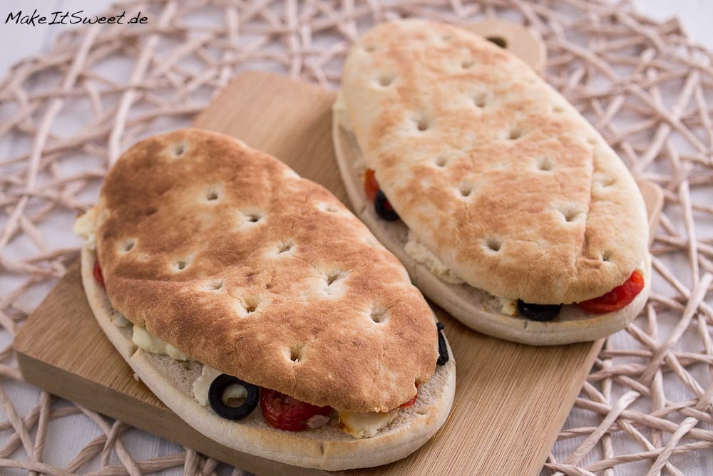 Zwei ovale Fladenbrote, belegt mit Feta, Oliven und Tomaten auf einem Holzbrett.