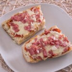 Pizza Baguette aus der Heißluftfritteuse Zwei Scheiben Baguette-Pizza, belegt mit geschmolzenem Käse, Schinken und gehackten Zwiebeln auf einem weißen Teller.