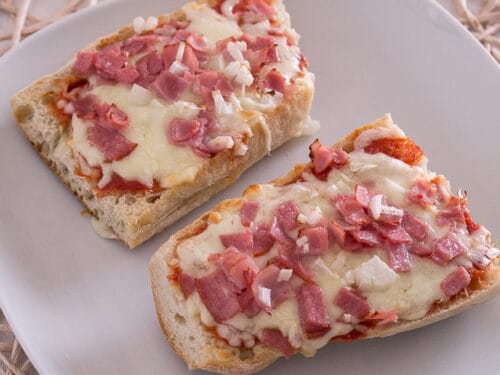 Zwei Scheiben Baguette-Pizza, belegt mit geschmolzenem Käse, Schinken und gehackten Zwiebeln auf einem weißen Teller.