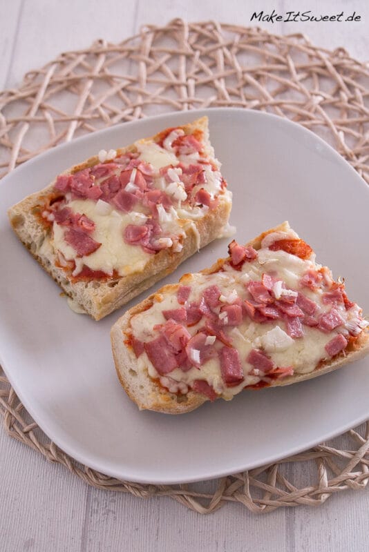 Zwei Scheiben Baguette-Pizza, belegt mit geschmolzenem Käse, Schinken und gehackten Zwiebeln auf einem weißen Teller.
