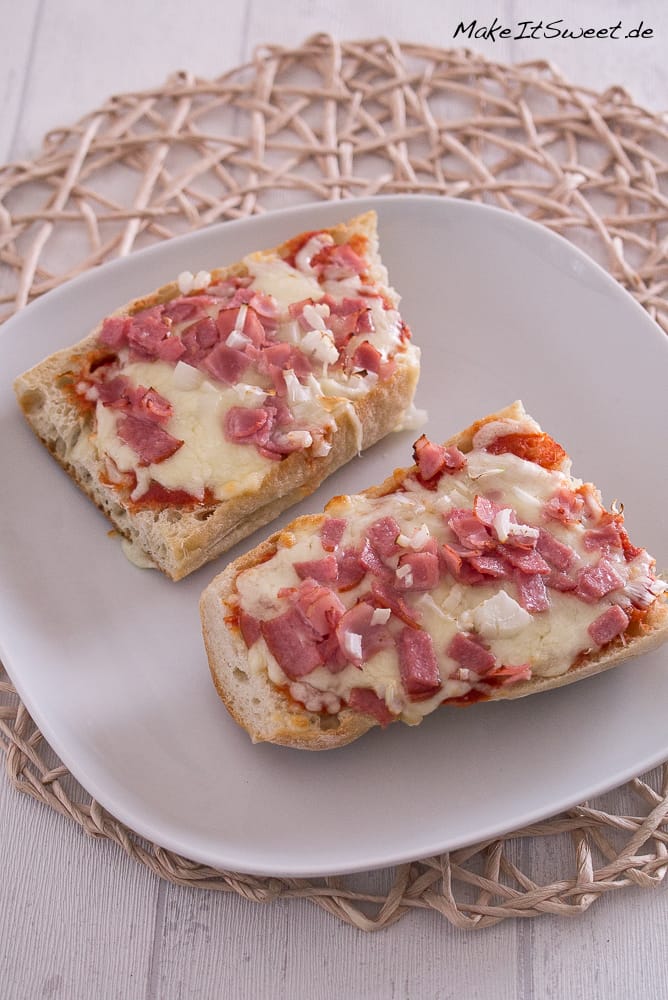 Pizza Baguette aus der Heißluftfritteuse 1 Zwei Scheiben Baguette-Pizza, belegt mit geschmolzenem Käse, Schinken und gehackten Zwiebeln auf einem weißen Teller.