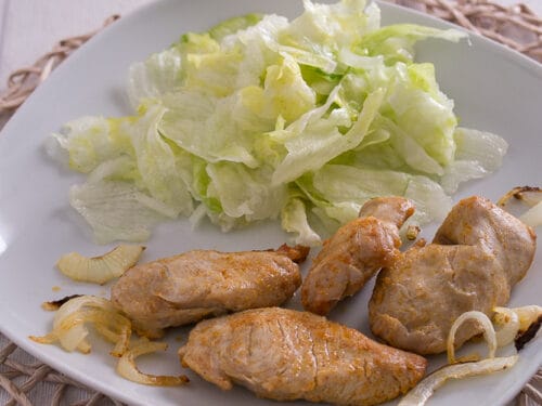 Gebratene Fleischstücke mit Zwiebeln und frischem Salat, serviert auf einem weißen Teller.