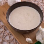 Schneller Schmand-Knoblauch-Dip Eine braune Schale mit cremigem Schmand-Dip, garniert mit Knoblauchzehen und -knollen, auf einem Holzbrett.