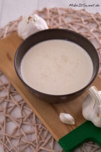 Eine braune Schale mit cremigem Schmand-Dip, garniert mit Knoblauchzehen und -knollen, auf einem Holzbrett.