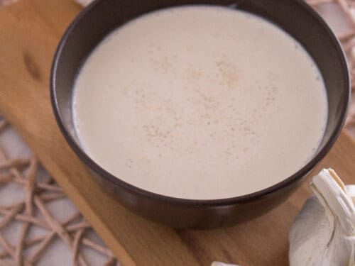 Eine braune Schale mit cremigem Schmand-Dip, garniert mit Knoblauchzehen und -knollen, auf einem Holzbrett.