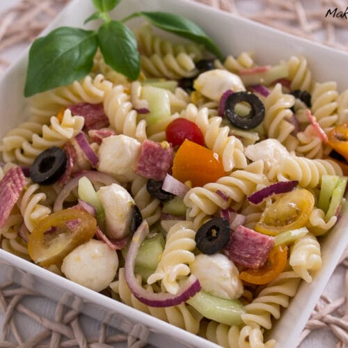 Nudelsalat Mozzarella, Oliven, Tomaten, Salami, Zwiebeln, Gurken und Basilikum in einer weißen Schüssel.