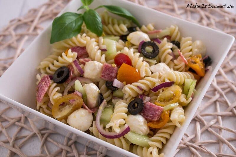Nudelsalat Mozzarella, Oliven, Tomaten, Salami, Zwiebeln, Gurken und Basilikum in einer weißen Schüssel.