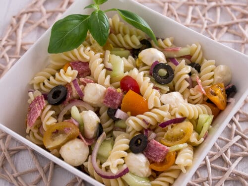 Italian Nudelsalat in einer quadratischen Schüssel mit Tomaten, Oliven, Mozzarella, Salami und Basilikum.