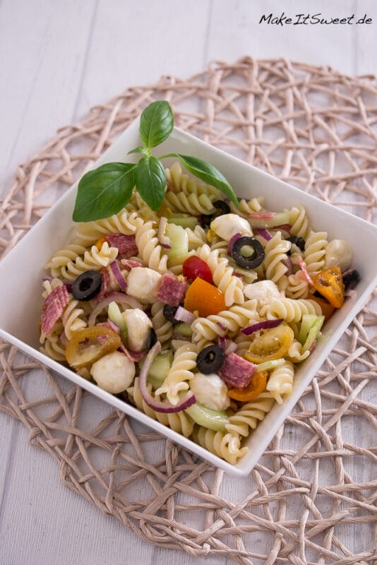 Italian Nudelsalat in einer quadratischen Schüssel mit Tomaten, Oliven, Mozzarella, Salami und Basilikum.