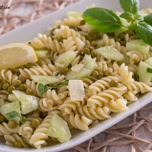 Nudelsalat mit Gurke, Erbsen, Käsewürfeln, Basilikumblättern und Zitronenscheiben auf einem weißen Teller.