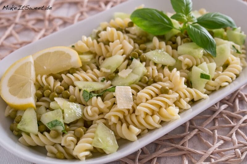 Nudelsalat mit Gurke, Erbsen, Käsewürfeln, Basilikumblättern und Zitronenscheiben auf einem weißen Teller.