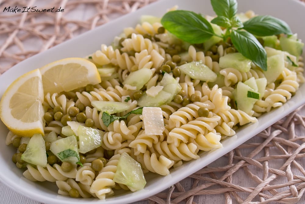 Nudelsalat mit Gurke, Erbsen, Käsewürfeln, Basilikumblättern und Zitronenscheiben auf einem weißen Teller.